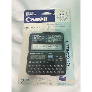 Może przedstawiać: Organizer Canon DM-320 w oryginalnym opakowaniu. Czarny organizer posiada klawiaturę, okno szybkiego podglądu i ekran. Opakowanie zawiera tekst "Przechowuje 164 imiona i nazwiska" oraz "Kalkulator 10-cyfrowy."