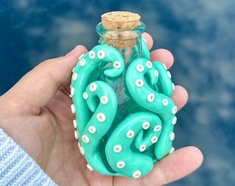 May include: A small glass bottle with a cork stopper is wrapped in a light green and white octopus-shaped clay sculpture. The octopus has white eyes and tentacles.