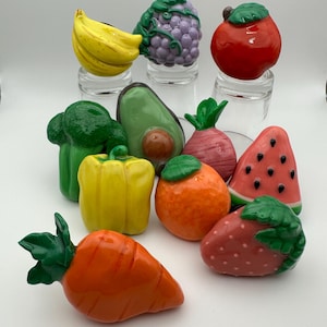 May include: A collection of ten colorful ceramic fruit and vegetable figurines. The figurines include a banana, grapes, apple, avocado, broccoli, bell pepper, radish, orange, watermelon, and strawberry.