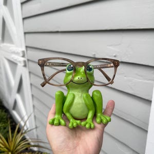 Op de afbeelding: Een groene kikkerfiguur met een bruin montuur bril. De bril wordt door de kikker vastgehouden. De bril is van Warby Parker.