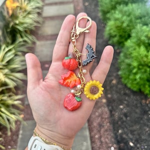 Op de afbeelding: Een goudkleurige sleutelhanger met meerdere bedels. Bedels zijn onder meer een rode pompoen, een herfstblad, een rode appel, een zwarte vleermuis en een gele zonnebloem. De sleutelhanger wordt in een hand gehouden.