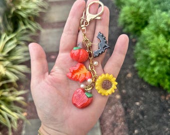 May include: A gold-colored keychain with multiple charms. Charms include a red pumpkin, a fall leaf, a red apple, a black bat, and a yellow sunflower. The keychain is held in a hand.