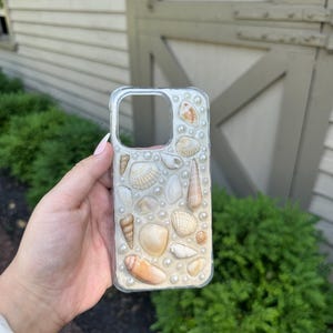 May include: A clear smartphone case decorated with an assortment of various tan, beige, and white seashells of different shapes and sizes, interspersed with small, round, white faux pearls embedded across the entire back surface of the cover.