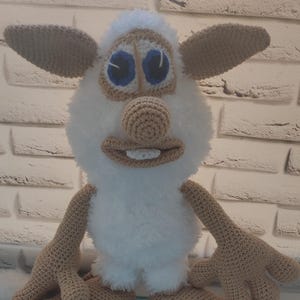 May include: A white crocheted plush toy with brown ears, a brown nose, and blue eyes. The toy has a toothy grin and is standing on a light blue wooden surface.