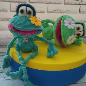 Può includere: Due peluche di rana verde all'uncinetto con occhi e bocche ricamate. Una rana è seduta con le gambe incrociate e ha un fiore giallo sulla testa. L'altra rana è sdraiata con la bocca aperta e ha fiori bianchi sulla testa. Entrambe le rane sono sedute su una piattaforma rotonda gialla e blu.