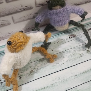 May include: Two handmade knitted frog toys. One frog is green and wearing a light purple sweater. The other frog is yellow and white. The toys are on a light wood surface with a brick wall background.