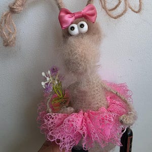 May include: A fuzzy, light brown creature figurine with long, thin, yarn-like ears, large white eyes, and a pink satin bow. It wears a bright pink lace tutu, pink plastic shoes, and holds a small bouquet of purple flowers and a miniature wine bottle.