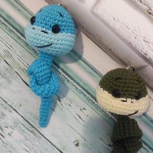 Pode incluir: Dois brinquedos de pelúcia de crochê em forma de pessoa. O primeiro brinquedo é azul e o segundo brinquedo é verde com o rosto branco.