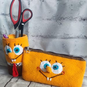 May include: A yellow crochet pencil holder and a matching rectangular crochet pencil case, both featuring the embroidered face of a cartoon character with large white eyes, blue irises, a nose, and teeth, with a star-shaped zipper pull.