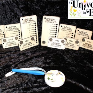 May include: A collection of wooden tags and a crochet hook with a blue handle. The tags feature the text "Universe in Bloom" and "2025". The crochet hook is white and blue. The background is a dark, textured fabric.