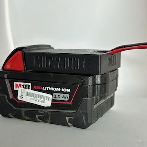 May include: Black M18 Red Lithium-Ion 3.0 Ah battery pack. The battery pack has the brand name "Milwaukee" embossed on the top. A red accent is visible on the side. A sticker with the text "ANW2085" is on the front.