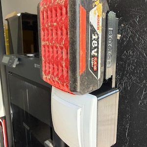 Op de afbeelding: Een rood en zwart Bosch 18V Li-ion batterijpakket is gemonteerd boven een wit en zwart elektrisch component. Het batterijpakket heeft een gestructureerd rood oppervlak en is bevestigd aan een zwarte muur.