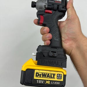 Puede incluir: Un atornillador de impacto Parkside negro y gris con un acento rojo, sostenido en una mano. El atornillador está conectado a una batería DeWalt de 18V amarilla y negra. La batería tiene el texto "4.0 Ah" y "18V/Li-Ion".