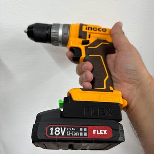 May include: A cordless drill with a black and yellow body, held in a hand. The drill has a black chuck and a black battery pack labeled "18V 2.5 Ah Li-Ion FLEX". The brand name "ingco" is visible on the drill body.