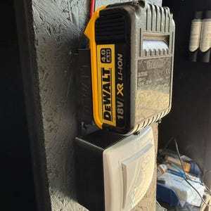 May include: A black and yellow DeWalt 18V 4.0 Ah Li-Ion battery pack attached to a black and white electrical switch. The battery pack is mounted on a textured, dark gray surface. A red wire extends from the battery.