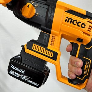 Può includere: Un trapano avvitatore arancione e nero senza fili con una batteria Makita da 18 V 3,0 Ah collegata. Il trapano ha il marchio "ingco" stampato sul lato e la parola "brushless" è visibile.