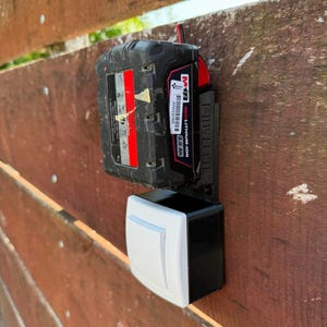 May include: A black and red M18 REDLITHIUM-ION 3.0 Ah battery attached to a black and white device, mounted on a brown wooden fence. The battery has a red stripe and white text.