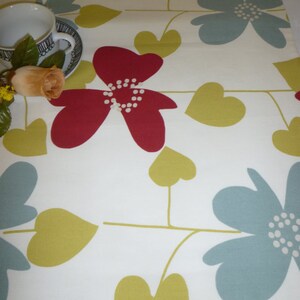 May include: Cream-coloured table runner with a floral design. It displays large red flowers with white centres, blue-green flowers, and yellow-green heart-shaped leaves on thin stems. A teacup and a rose are in the upper left corner.