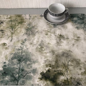 May include: A white table runner with a green and grey watercolour forest print. The runner features a blurred, abstract design of trees and foliage.