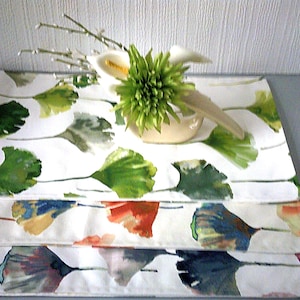 May include: Three white placemats featuring a watercolour ginkgo leaf design in green, blue, and orange hues. A small vase holds a green floral arrangement and white calla lilies. The placemats are stacked on a grey surface.