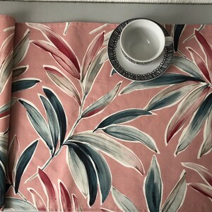 May include: A pink table runner featuring a botanical leaf design in red, blue-green, and grey. A white teacup and saucer with a black patterned rim are placed on the runner. The runner is ideal for adding a decorative touch to a table.