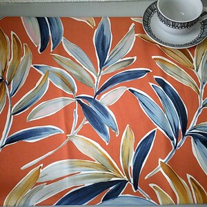 May include: An orange table runner featuring a botanical leaf design in blue, white, and tan. A teacup and saucer with a black and white pattern are placed on the runner. The runner is likely made of fabric.