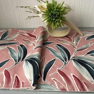 May include: Pink fabric featuring a botanical leaf design in blue, grey, and red. A decorative floral arrangement with white calla lilies and a green accent sits on the fabric.
