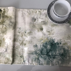 May include: A white table runner with a green and grey abstract design. The design features a watercolour-like effect with branches and leaves.