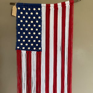 May include: Macrame wall hanging depicting the American flag. The blue field features white knots representing stars. Red and white cords form the stripes. The flag is suspended from a natural wood branch.