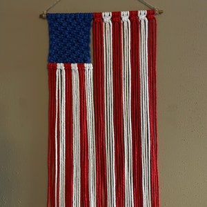 May include: A red, white, and blue macrame wall hanging in the shape of the American flag. The flag is made with yarn and has a wooden dowel at the top.