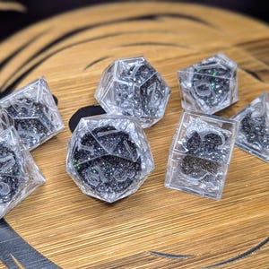 May include: A collection of seven clear resin dice, each filled with black glitter and white numerals. The dice, in shapes like d4, d6, d8, d10, d12, and d20, are displayed on a wooden surface.