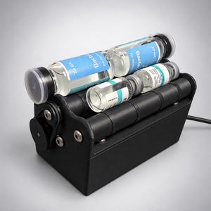 May include: A black laboratory roller mixer device featuring a series of horizontal rotating cylinders. Two clear glass vials with blue labels marked 10 mL and the text Bacteriostatic Water for Injection are positioned on top of the rollers.
