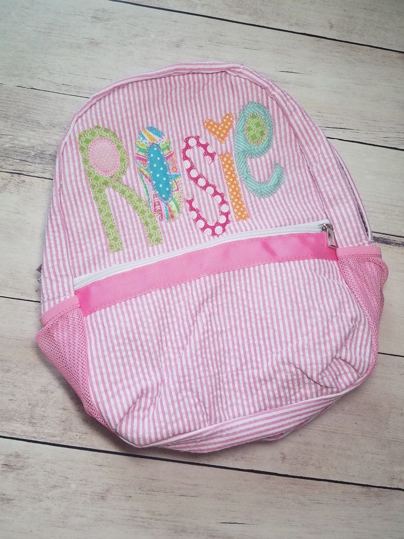 May include: Pink and white striped backpack with the name "Rosie" embroidered on the front in colorful fabric letters.