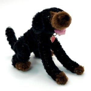 Puede incluir: Figura de perro negro y marrón hecha a mano con limpiapipas difusos. El perro tiene una lengua rosa, una etiqueta roja en forma de corazón y patas marrones. El perro está en una pose juguetona.