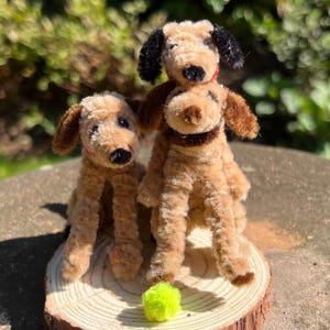 May include: Three brown pipe cleaner dogs, one on top of the other, with a green ball in front of them. The dogs are sitting on a wooden slice.