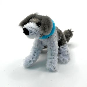 May include: A handmade pipe cleaner dog figurine in shades of gray and white, with a black nose and a bright blue collar. The dog is posed in a playful stance, with its tail slightly raised. The craft is set against a plain white background.