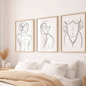以下が含まれることがあります： 女性の姿を描いた3つの額入り線画プリント。ミニマリストな黒い線画は、明るい木製のフレームに入った白い背景にあります。寝室は、白い寝具とベージュのスローを備えたニュートラルなカラーパレットです。