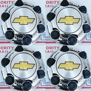 May include: A set of four circular chrome wheel center caps, each featuring a prominent gold Chevrolet bowtie emblem in the center and six black lug nut covers arranged around the perimeter of the polished metallic surface.