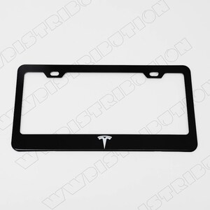 May include: Black license plate frame with the Tesla logo in white. The frame is rectangular with rounded corners and two mounting holes at the top. The Tesla logo is centered at the bottom.