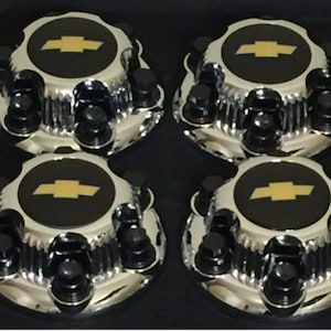 May include: A set of four circular chrome wheel center caps featuring a black center disc with a gold Chevrolet bowtie emblem. Each cap has a fluted outer edge and is accented with eight black lug nut covers arranged in a circular pattern.