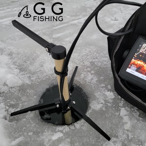May include: A black ice fishing rod holder with a wooden pole and three adjustable legs. The holder is set up on a frozen lake with a fish finder in a black bag nearby. The logo "GG FISHING" is visible in the top left corner of the image.