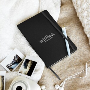 Può includere: Un diario nero con copertina rigida con il testo "2021 selfmade design your life" sulla copertina. Il diario è appoggiato su una superficie soffice bianca e marrone con una penna blu e un paio di auricolari bianchi.