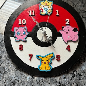 May include: A black and white clock with red accents features a Pokeball design. The clock face has numbers 1 through 12 and features three Pokemon characters: Pikachu, Jigglypuff, and  a yellow and white Pokemon with a crown.