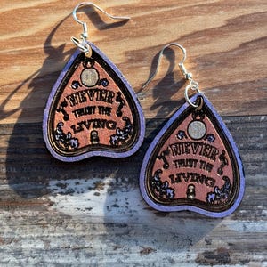 May include: A pair of earrings shaped like planchette boards, with the text "NEVER TRUST THE LIVING". The earrings have a purple border, a copper-colored center, and silver-colored hooks. The earrings are made of wood.