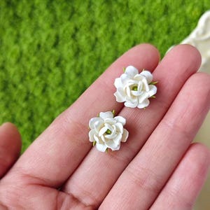 Op de afbeelding: Twee witte bloemvormige oorstekers met groene centra en groene accenten op de bloemblaadjes. De oorbellen worden in een hand gehouden tegen een groene achtergrond. De oorbellen zijn klein en delicaat.