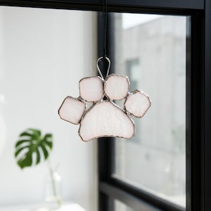 May include: A stained glass suncatcher shaped like a paw print, featuring translucent pale pink glass segments held together by silver-toned metal solder lines, suspended from a thin black cord against a window pane.