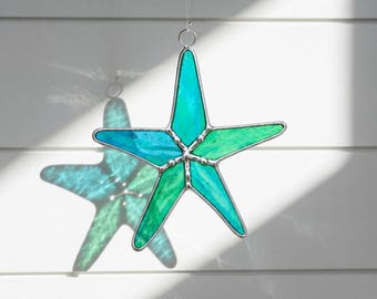 May include: A five-pointed starfish suncatcher crafted from translucent stained glass in varying shades of teal, turquoise, and emerald green. The glass segments are joined by thin silver-toned metal solder lines and include a small metal ring at the top.