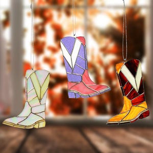 May include: Three stained glass cowboy boot ornaments in shades of yellow, purple, red, and white. Each boot has a silver string for hanging. The boots are designed with geometric shapes and are suitable for home decor.