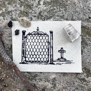 Può includere: Una piccola toppa di tessuto rettangolare con un'illustrazione in bianco e nero di un cancello e una croce. Un ditale trasparente e un pezzo di pigna sono accanto. Su una superficie grigia strutturata.