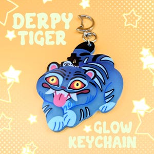 Puede incluir: Un llavero de tigre azul con un cierre de metal plateado. El tigre tiene ojos amarillos, lengua rosa y colmillos blancos. El texto DERPY TIGER y GLOW KEYCHAIN es visible. El tigre presenta rayas azul oscuro y un pequeño sombrero negro.